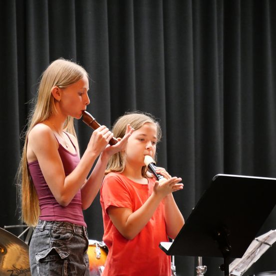 Zwei junge Mädchen spielen Blockflöten in einem Musikraum, wobei eines ein lila Tanktop und das andere ein orangefarbenes T-Shirt trägt. Sie stehen vor einem schwarzen Vorhang und einem Notenständer.