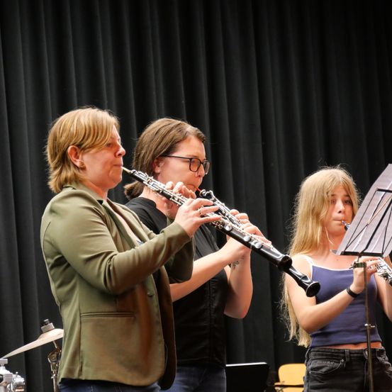 Drei Frauen spielen in einem Raum Musikinstrumente. Die Frau auf der linken Seite spielt Klarinette, die Frau in der Mitte Flöte und die Frau auf der rechten Seite Trompete. Sie tragen alle Freizeitkleidung und stehen vor einem schwarzen Vorhang.