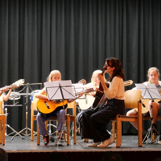 Eine Gruppe junger Mädchen und eine Frau spielen auf der Bühne Gitarre. Die Frau sitzt, während die Mädchen stehen. Sie alle schauen auf ihre Notenblätter.