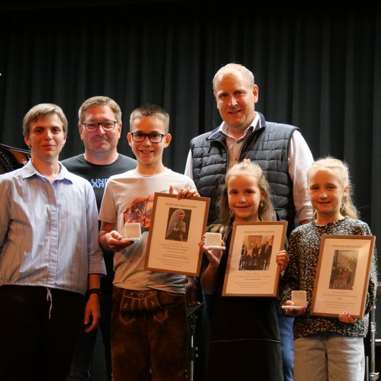 Fünf Personen, darunter drei Kinder, stehen zusammen und halten gerahmte Urkunden, lächeln für ein Foto auf einer Bühne.
