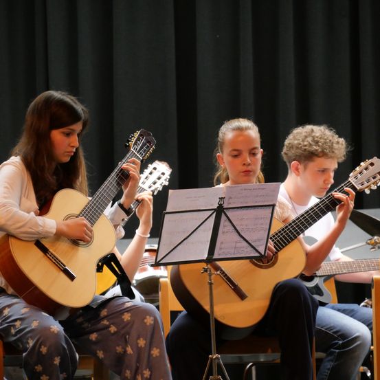 Drei junge Musiker, zwei Mädchen und ein Junge, spielen Gitarren in einem Klassenzimmer. Das Mädchen links konzentriert sich auf ihre Gitarre, während die anderen beiden Notenblätter betrachten.