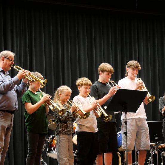 Eine Gruppe junger Musiker, darunter Jungen und Mädchen, spielen Trompeten auf der Bühne, begleitet von einem Lehrer. Sie treten bei einem Schulkonzert auf.