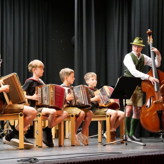 Fünf junge Jungen sitzen auf der Bühne, jeder spielt ein Akkordeon, begleitet von einem Mann, der Cello spielt.