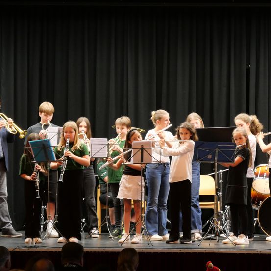 Eine Gruppe von Kindern auf der Bühne, die verschiedene Musikinstrumente spielen, darunter Flöten, Trompeten und Trommeln, unter einem schwarzen Vorhang.