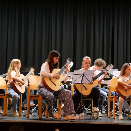 Eine Gruppe junger Mädchen spielt auf der Bühne mit Gitarren, mit einem schwarzen Vorhang als Hintergrund.