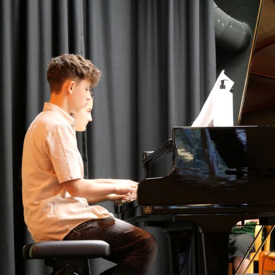 Ein junger Junge spielt Klavier, während ein anderer Junge ihm genau zusieht. Das Klavier ist schwarz, und sie stehen vor einem schwarzen Vorhang.