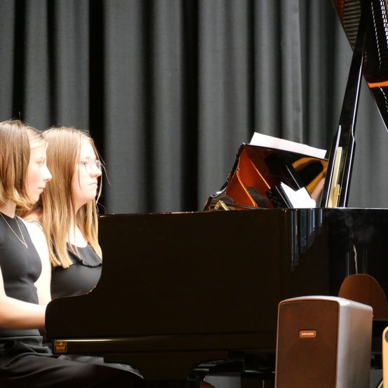 Zwei junge Mädchen in schwarzen Kleidern beobachten aufmerksam eine Klavieraufführung auf der Bühne. Das Klavier ist schwarz mit einem Lautsprecher in der Nähe.