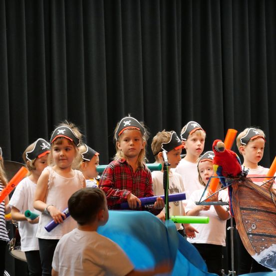 Eine Gruppe von Kindern, die als Piraten gekleidet sind, halten Musikinstrumente in der Hand und stehen auf einer Bühne mit einem Piratenschiff als Hintergrund.