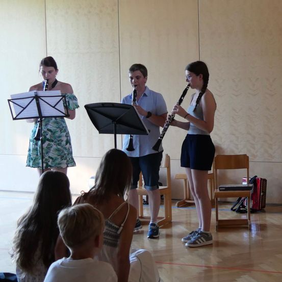 Drei junge Musiker, zwei Mädchen und ein Junge, spielen Klarinetten in einem Raum mit Holzböden und -wänden. Sie stehen vor Notenständern mit Notenblättern. Ein paar Kinder sitzen vor ihnen auf dem Boden.