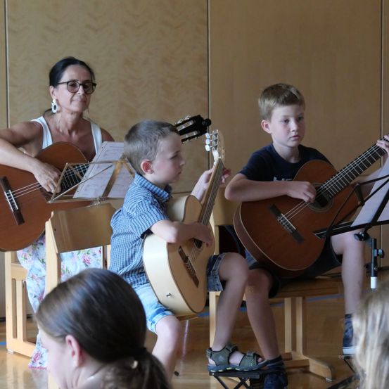 Eine Frau und zwei Jungen spielen Gitarre in einem Raum. Die Frau sitzt auf einem Stuhl mit einem Notenständer, und die Jungen sitzen vor ihr auf Stühlen.