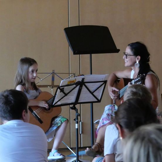 Zwei Frauen, eine junges Mädchen und eine Erwachsene, spielen Gitarre vor einem Publikum. Das Mädchen sitzt und trägt einen Blumenrock, während die Frau steht und eine Brille trägt.