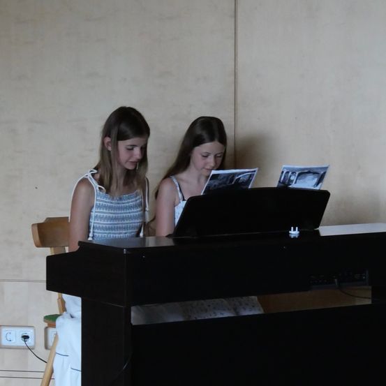 Zwei junge Mädchen sitzen an einem Klavier, eine liest Noten, die andere konzentriert sich auf die Tasten. Das Klavier ist schwarz, und sie befinden sich in einem Raum mit einer beigen Wand.