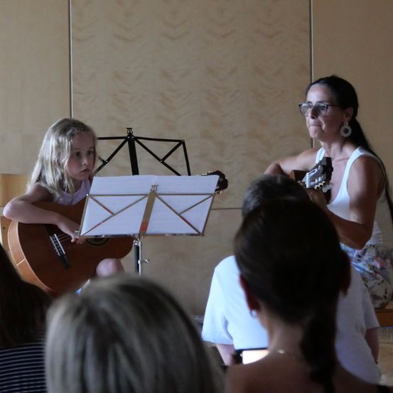 Ein junges Mädchen spielt Gitarre und liest Noten, begleitet von einer erwachsenen Frau, vor einem sitzenden Publikum.