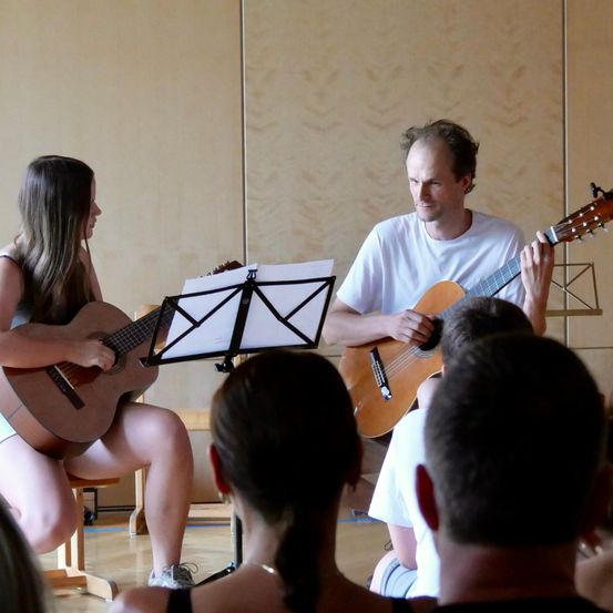 Ein Mann und eine Frau spielen Gitarre vor einer Gruppe von Menschen. Die Frau sitzt auf einem Stuhl, und der Mann steht. Sie schauen beide auf das Notenblatt auf dem Notenständer.