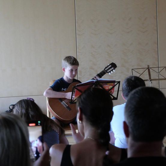 Ein junger Junge spielt Gitarre vor einem kleinen Publikum, mit einem Notenständer vor ihm.