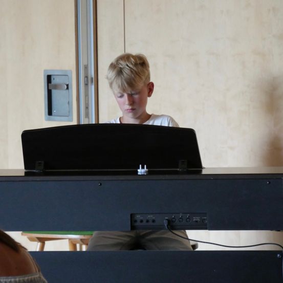 Ein junger Junge sitzt an einem Klavier und konzentriert sich auf das vor ihm liegende Notenblatt. Das Klavier ist schwarz mit einem digitalen Display, und er trägt ein weißes Hemd.