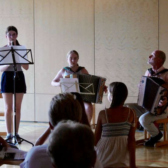 Eine Gruppe von Musikern tritt auf der Bühne auf, während ein Publikum zusieht. Eine Frau spielt Flöte, eine andere spielt Akkordeon und ein Mann spielt Akkordeon.