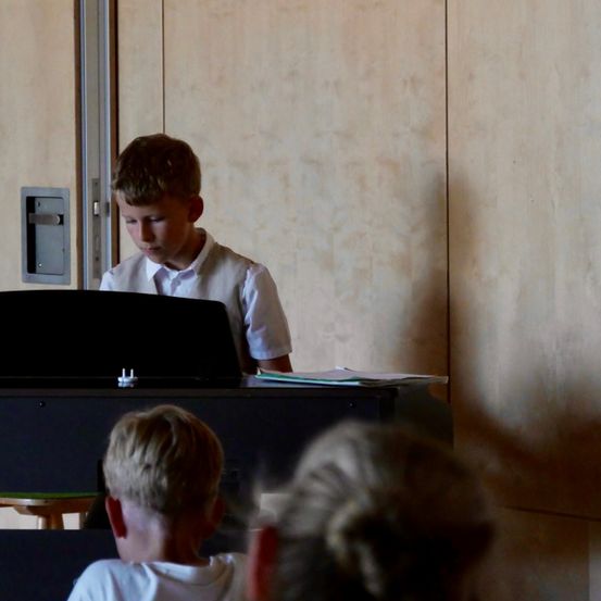 Ein junger Junge in einem weißen Hemd spielt Klavier, während ein anderer Junge und eine Frau von hinten zusehen.