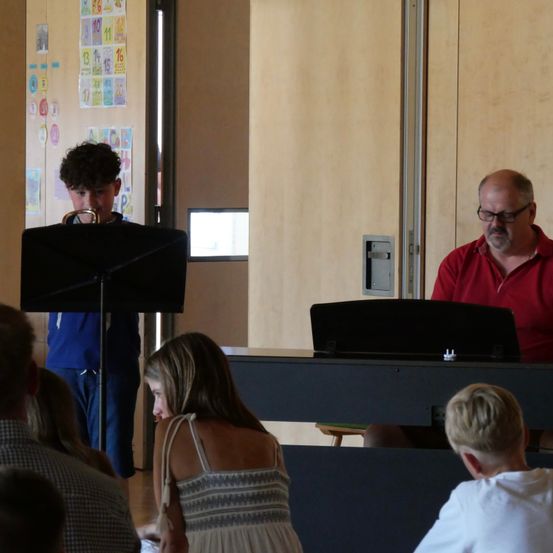 Ein Junge spielt Trompete, während ein Mann in einem roten Hemd Klavier spielt. Eine Frau und ein Junge schauen von vorne zu.