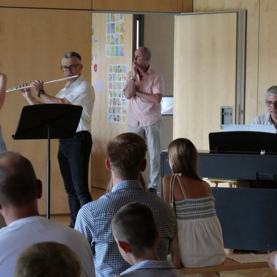 Ein Mann spielt Flöte, während ein anderer Mann in ein Mikrofon spricht in einem Klassenzimmer. Eine Frau spielt Klavier im Hintergrund.