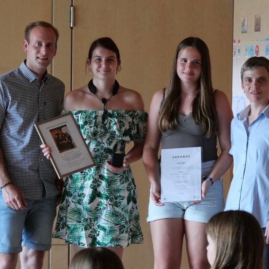 Vier Personen stehen in einem Raum, zwei Frauen halten Urkunden und ein gerahmtes Bild, während ein Mann neben ihnen lächelt.