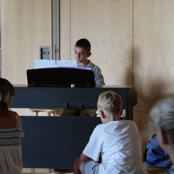 Ein junger Junge spielt Klavier, während eine Gruppe von Kindern und Erwachsenen aufmerksam zusieht.