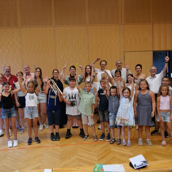 Eine Gruppe von Erwachsenen und Kindern, einige mit Musikinstrumenten, posiert für ein Foto auf einer Bühne mit Holzboden.