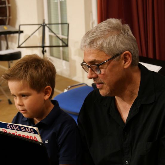 Ein älterer Mann mit Brille schaut einen jungen Jungen an, der ein Musikbuch mit dem Titel Jessie Blake in der Hand hält.