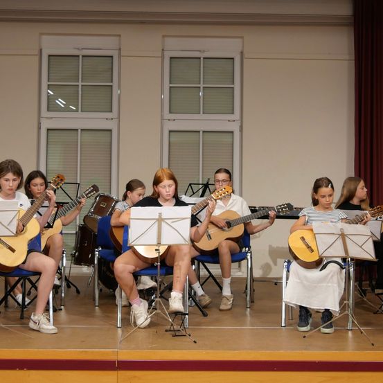 Eine Gruppe junger Kinder sitzt auf Stühlen auf der Bühne und spielt Gitarre und Schlagzeug. Sie schauen in ihre Notenblätter.