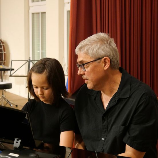 Ein Mann und ein junges Mädchen sitzen gemeinsam am Klavier, der Mann trägt eine Brille und das Mädchen konzentriert sich auf das Notenblatt.