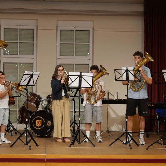 Fünf junge Musiker spielen Blechblasinstrumente auf einer Bühne, einer spielt Trompete, zwei spielen Posaunen und zwei spielen Tubas. Sie werden von einem Schlagzeug und einem Klavier begleitet.