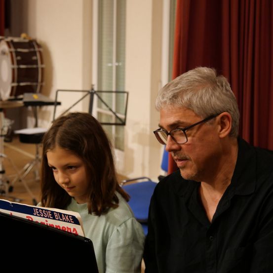 Ein Mann mit Brille und schwarzem Hemd schaut sich mit einem jungen Mädchen in einem grünen Shirt eine Musikpartitur an. Dahinter steht ein Schlagzeug und ein Notenständer.