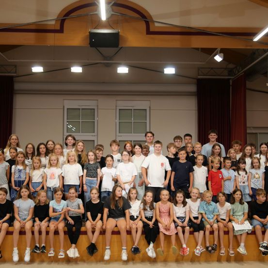 Eine Gruppe von Kindern und Jugendlichen sitzt und steht auf einer Bühne und posiert für ein Foto. Sie sind lässig gekleidet und einige tragen Brillen. Die Bühne hat eine Holzplattform und eine weiße Wand mit zwei Fenstern.