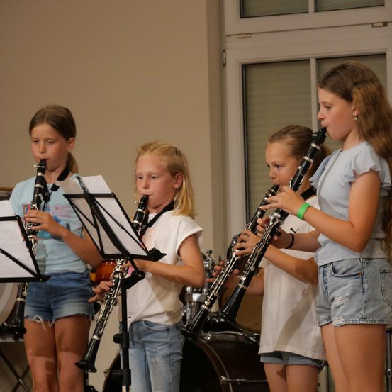 Vier junge Mädchen spielen Klarinetten in einem Raum mit einem Schlagzeug im Hintergrund.