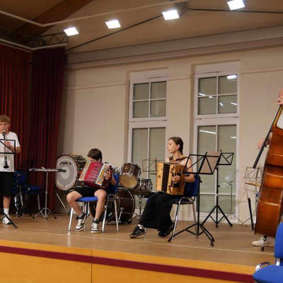 Eine Gruppe Musiker tritt auf der Bühne auf, wobei zwei Personen sitzend Akkordeons spielen, eine Person steht und Klarinette spielt und eine andere Cello spielt. Die Bühne ist gut beleuchtet, und das Publikum ist nicht sichtbar.