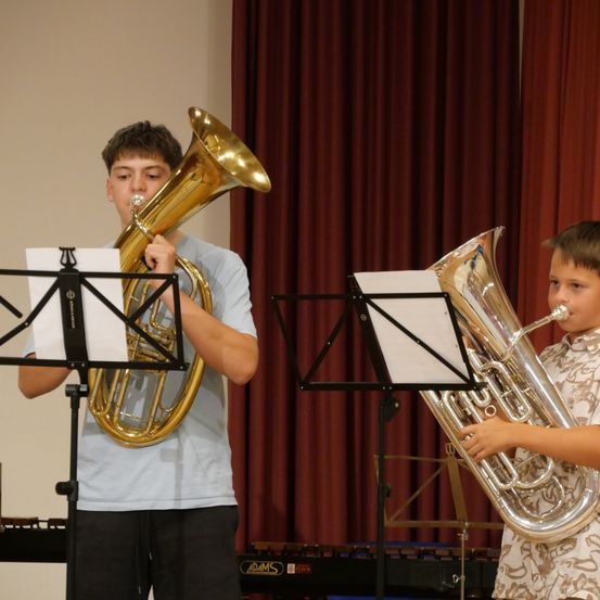 Zwei junge Jungen spielen Tubas in einem Raum mit roten Vorhängen und einem Klavier im Hintergrund.