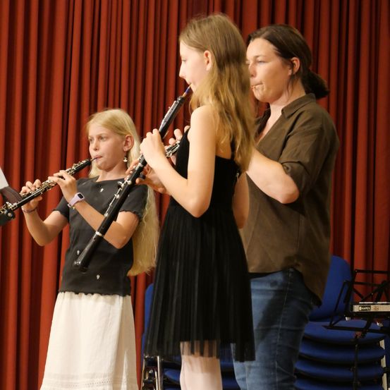 Zwei junge Mädchen spielen Klarinetten, während eine Frau, möglicherweise ihre Lehrerin, hinter ihnen steht, vor einem roten Vorhang.