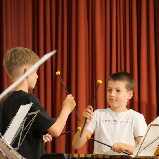 Zwei Jungen spielen Xylophon in einer Aufführung. Einer trägt ein schwarzes T-Shirt, der andere ein weißes mit Tommy Hilfiger-Logo. Sie konzentrieren sich auf ihre Instrumente.
