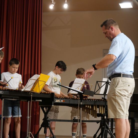 Ein Mann unterrichtet eine Gruppe von Jungen, die Xylophon spielen, in einem Raum mit einem roten Vorhang und einem Klavier im Hintergrund.
