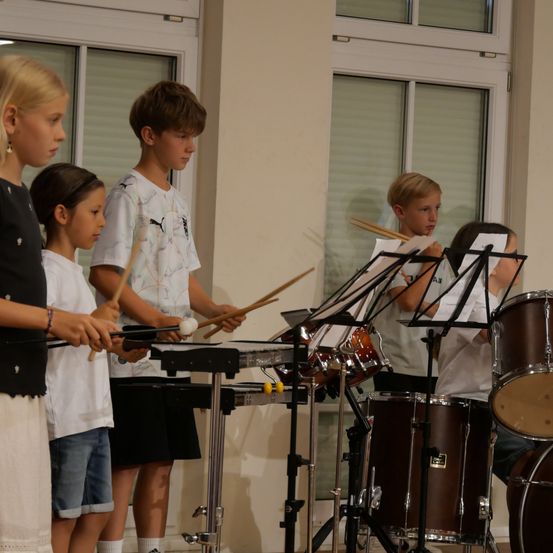 Fünf Kinder spielen in einem Raum mit Glasfenstern Instrumente. Ein Mädchen links spielt Xylophon, während der Junge neben ihr Schlagzeug spielt. Ein anderer Junge spielt Flöte. Ein Mädchen spielt Schlagzeug.