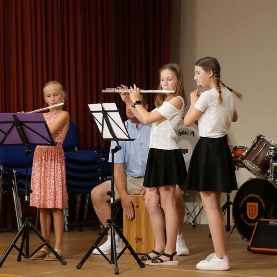 Drei junge Mädchen spielen Flöten auf einer Bühne. Ein Mann sitzt hinter ihnen und schaut zu. Musikinstrumente und Stühle sind im Hintergrund.