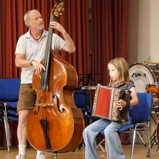 Ein älterer Mann spielt ein großes Cello, während ein junges Mädchen auf einem Stuhl sitzt und ein Akkordeon spielt. Sie sind in einem Raum mit roten Vorhängen und Musikinstrumenten im Hintergrund.