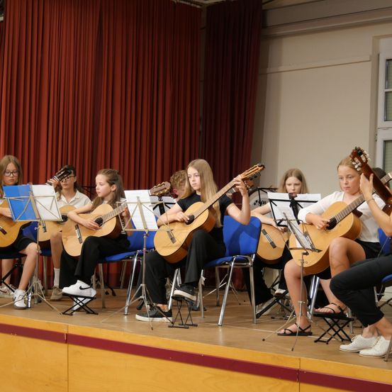 Eine Gruppe junger Mädchen spielt Gitarren auf der Bühne, mit roten Vorhängen im Hintergrund und Notenständern vor ihnen.