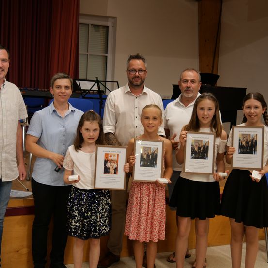 Eine Gruppe von Erwachsenen und Kindern steht zusammen und hält Urkunden in der Hand, lächelt für ein Foto in einem Raum mit einer Holzwand und einem Klavier im Hintergrund.