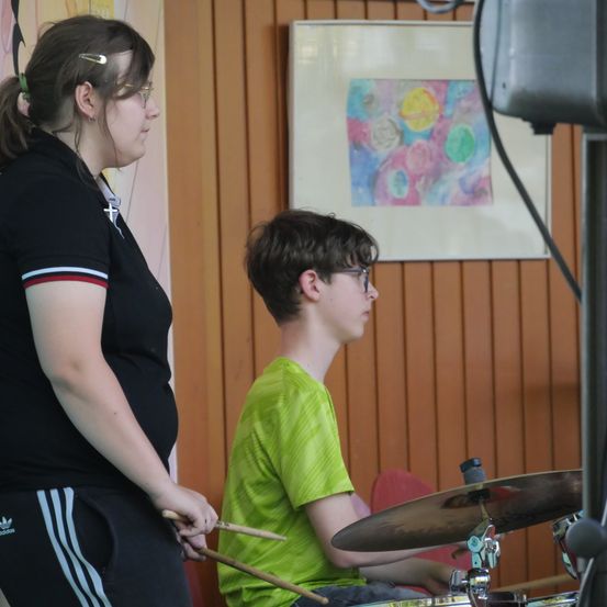 Eine Frau in einem schwarzen Shirt steht neben einem Jungen in einem grünen Shirt, beide halten Drumsticks, mit einem Schlagzeug vor ihnen und einem farbenfrohen Gemälde an der Wand dahinter.