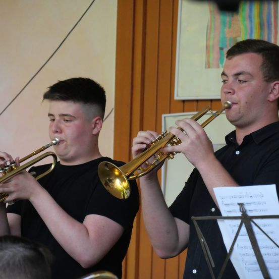 Zwei junge Männer spielen Trompeten in einem Raum, einer hält ein Musikblatt. Sie sind in schwarzen Hemden gekleidet.