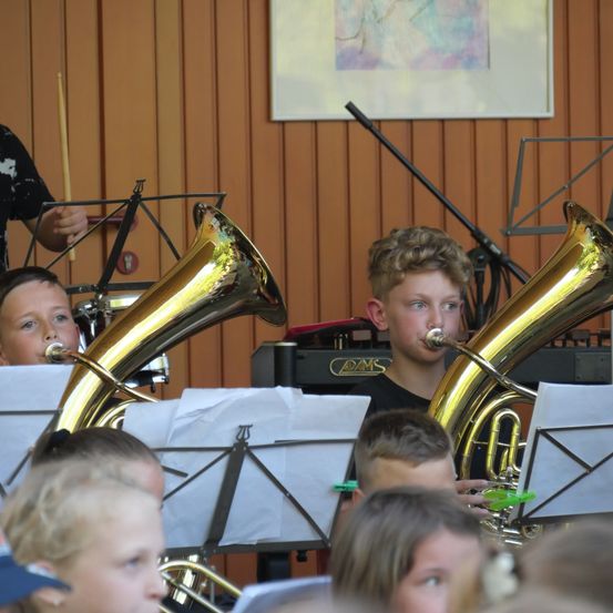 Eine Gruppe von Kindern spielt Blechblasinstrumente, einschließlich Tubas, in einem Musikraum. Sie sind von Notenständern und einer Wand mit einem gerahmten Bild umgeben.