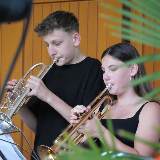 Zwei junge Musiker, ein Junge und ein Mädchen, spielen Trompeten in einem Raum mit Holzwänden und grünen Pflanzen.