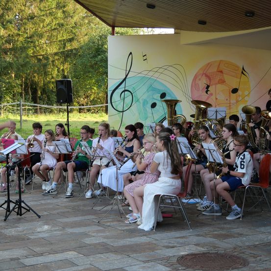 Eine Gruppe von Kindern in einer Band spielt Instrumente in einer Außenumgebung mit einem Wandbild im Hintergrund.