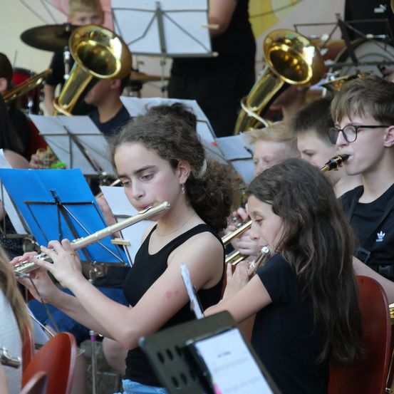 Eine Gruppe junger Musiker, darunter Mädchen, die Flöten spielen, tritt in einer Schulband auf. Die Band sitzt im Halbkreis, mit Notenständern vor ihnen.